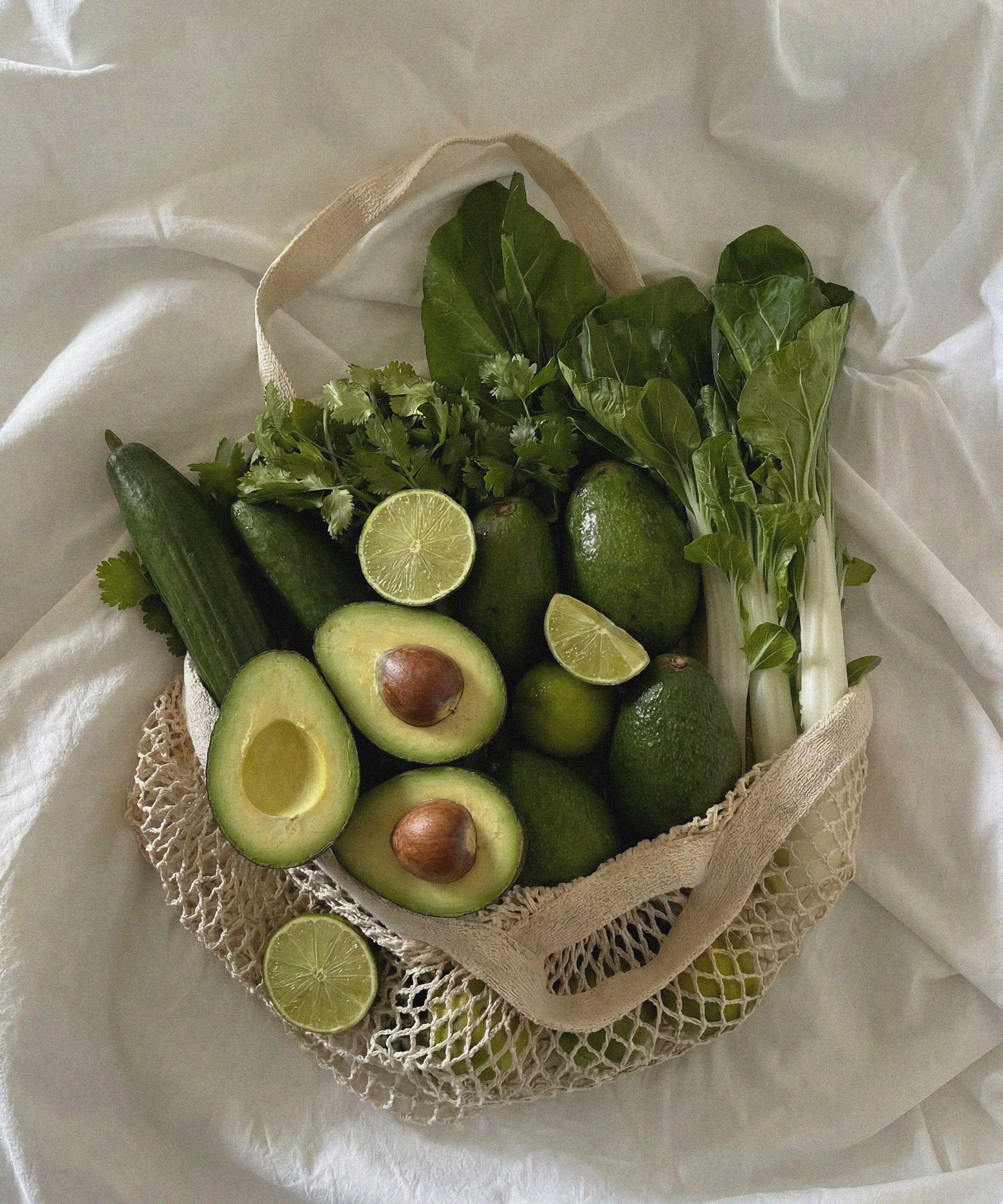 Mesh bag filled with green fruits and vegetables on a white fabric background